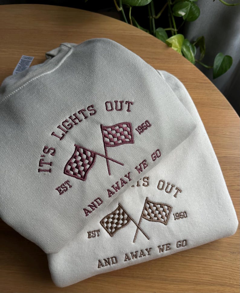 K&ouml;nnte beinhalten: Ein hellgraues Sweatshirt mit dem Text "IT'S LIGHTS OUT EST 1950 AND AWAY WE GO EST 1950 AND AWAY WE GO", der in Maroon gestickt ist. Das Sweatshirt zeigt zwei gekreuzte karierte Flaggen, eine in Maroon und eine in Braun.