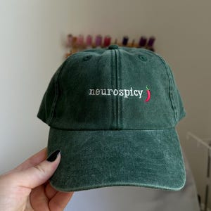 Op de afbeelding: Een donkergroene baseballpet met een gewassen, vintage textuur. De voorkant is voorzien van de wit geborduurde tekst "neurospicy" gevolgd door een klein, rood chilipeper-icoontje met een groen steeltje, gecentreerd boven de gebogen klep.