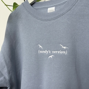 May include: A light blue sweatshirt with white embroidered birds and the text "(sandy's version)"
