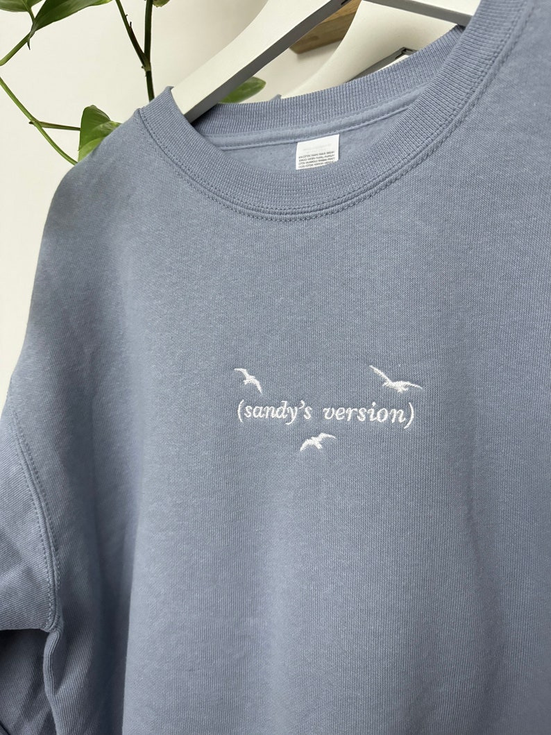 May include: A light blue sweatshirt with white embroidered birds and the text "(sandy's version)"