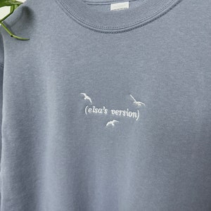 May include: A light blue sweatshirt with white embroidered birds and the text "(elsa's version)"