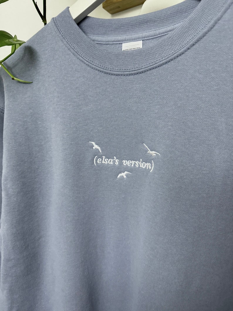 May include: A light blue sweatshirt with white embroidered birds and the text "(elsa's version)"