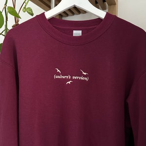 May include: Burgundy sweatshirt with a white embroidered design of birds flying and the text "(auburn's version)"