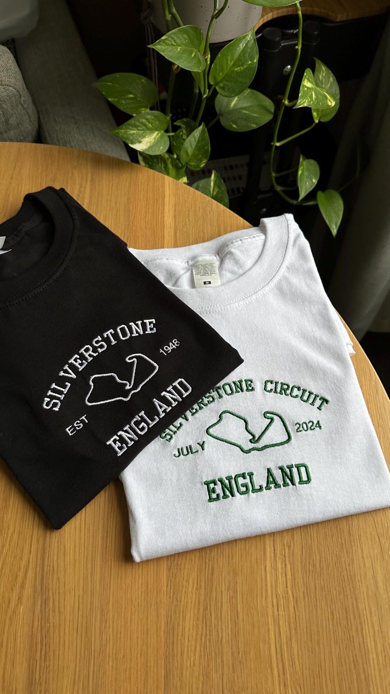 Pode incluir: Uma t-shirt preta com bordado branco com a inscri&ccedil;&atilde;o SILVERSTONE 1948 EST ENGLAND e o contorno de um circuito, e uma t-shirt branca com bordado verde com a inscri&ccedil;&atilde;o SILVERSTONE CIRCUIT JULY 2024 ENGLAND e o contorno de um circuito, ambas com gola redonda.