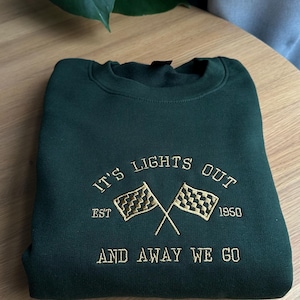 May include: Green sweatshirt with brass embroidered text that reads "It's Lights Out Est. 1950 And Away We Go" with a chequered flag graphic.