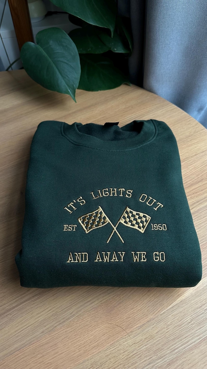 May include: Green sweatshirt with brass embroidered text that reads "It's Lights Out Est. 1950 And Away We Go" with a chequered flag graphic.