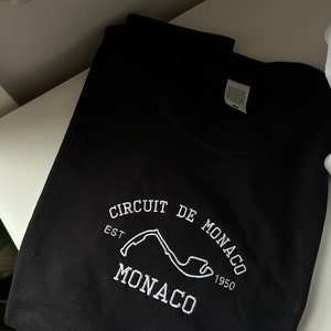 Pode incluir: Uma t-shirt preta com um bordado branco do tra&ccedil;ado do circuito de M&oacute;naco. O texto "CIRCUIT DE MONACO" aparece em arco sobre o circuito, com "EST 1950" nos lados e a palavra "MONACO" bordada em letras grandes na parte inferior.