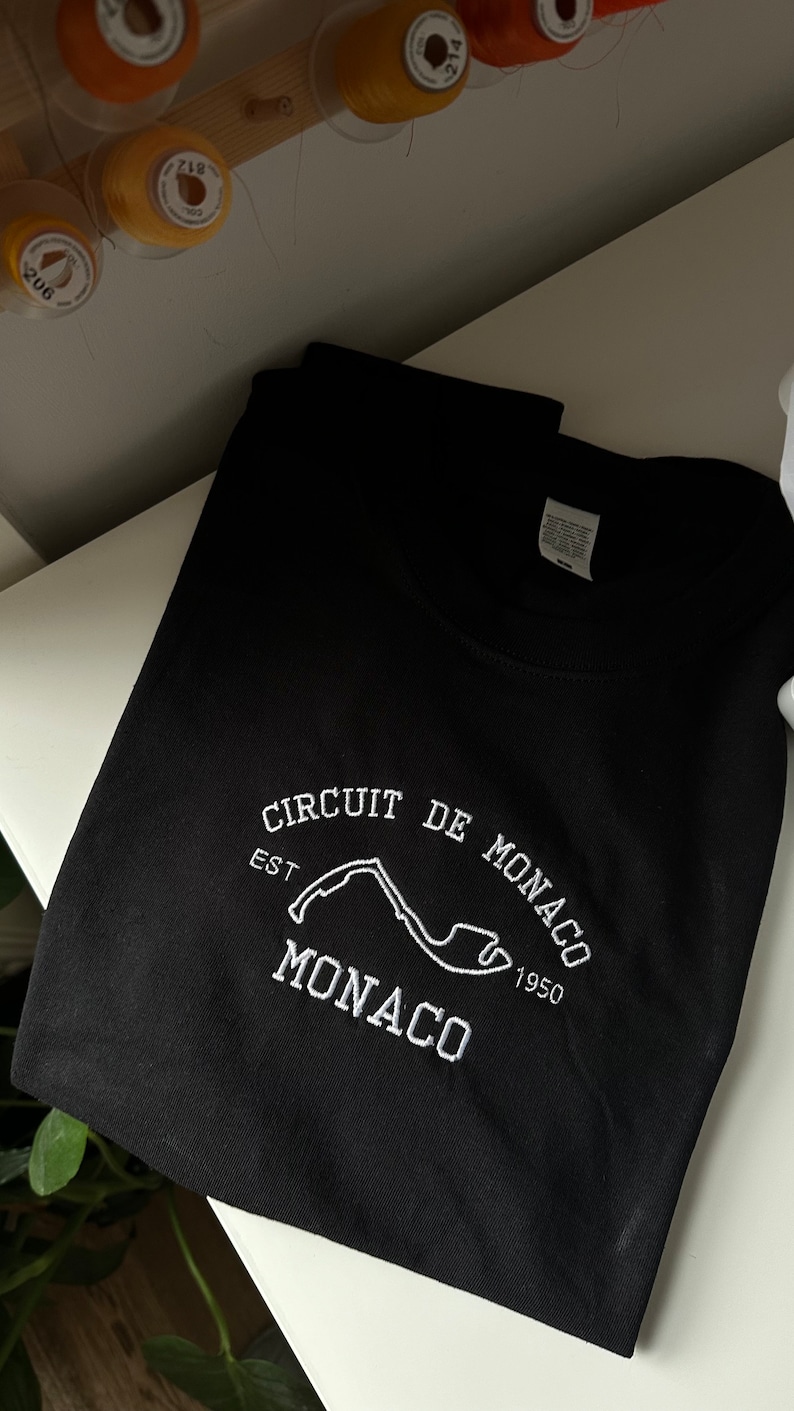 Pode incluir: Uma t-shirt preta com um bordado branco do tra&ccedil;ado do circuito de M&oacute;naco. O texto "CIRCUIT DE MONACO" aparece em arco sobre o circuito, com "EST 1950" nos lados e a palavra "MONACO" bordada em letras grandes na parte inferior.