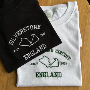 Pode incluir: Uma t-shirt preta com bordado branco com a inscri&ccedil;&atilde;o SILVERSTONE EST 1948 ENGLAND e o contorno de um circuito, e uma t-shirt branca com bordado verde com a inscri&ccedil;&atilde;o SILVERSTONE CIRCUIT JULY 2024 ENGLAND e o contorno de um circuito, ambas com gola redonda.