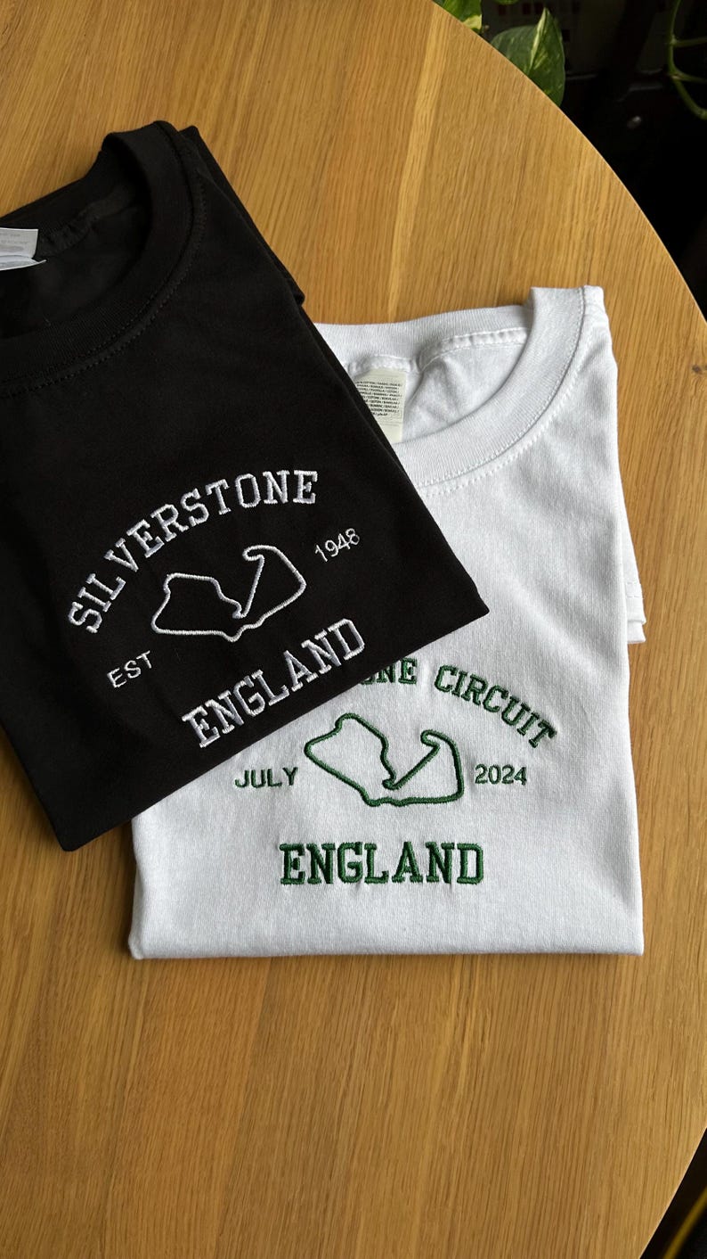 Pode incluir: Uma t-shirt preta com bordado branco com a inscri&ccedil;&atilde;o SILVERSTONE EST 1948 ENGLAND e o contorno de um circuito, e uma t-shirt branca com bordado verde com a inscri&ccedil;&atilde;o SILVERSTONE CIRCUIT JULY 2024 ENGLAND e o contorno de um circuito, ambas com gola redonda.