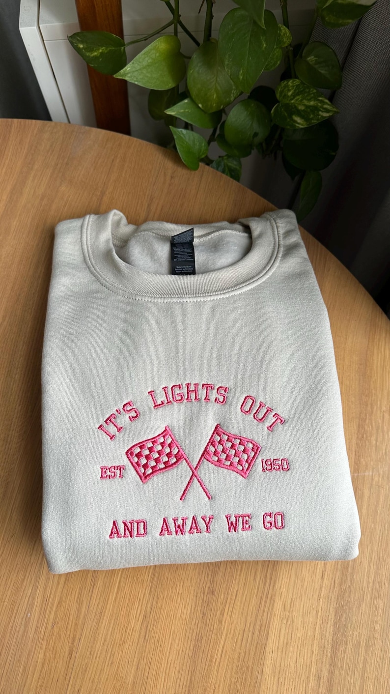 May include: A sand sweatshirt with pink embroidered text that reads "It's Lights Out Est. 1950 And Away We Go" with a chequered flag graphic.