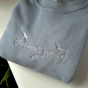 May include: A light blue sweatshirt with white embroidery that reads "(Rebecca's version)" and features two white embroidered birds.
