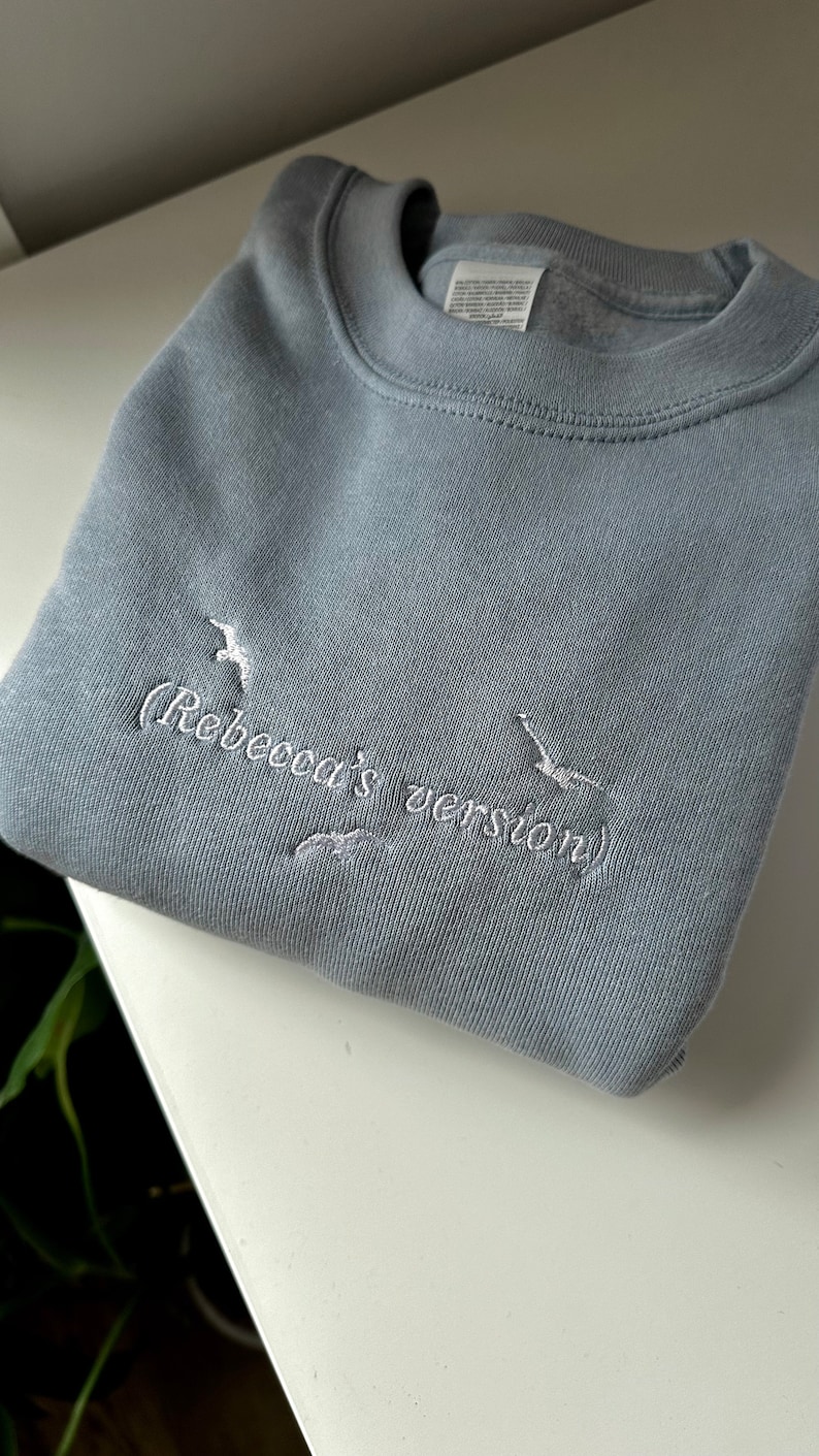 May include: A light blue sweatshirt with white embroidery that reads "(Rebecca's version)" and features two white embroidered birds.