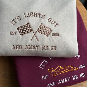 K&ouml;nnte beinhalten: Zwei Kapuzenpullover, einer beige und einer burgunderrot, mit gestickten Designs zum Thema Rennsport. Der beige Pullover hat ein kariertes Flaggen-Design und den Text "IT'S LIGHTS OUT EST 1950 AND AWAY WE GO". Der burgunderrote Pullover hat ein stilisiertes Rennwagen-Design und den Text "IT'S EST 1950 AND AWAY WE GO".