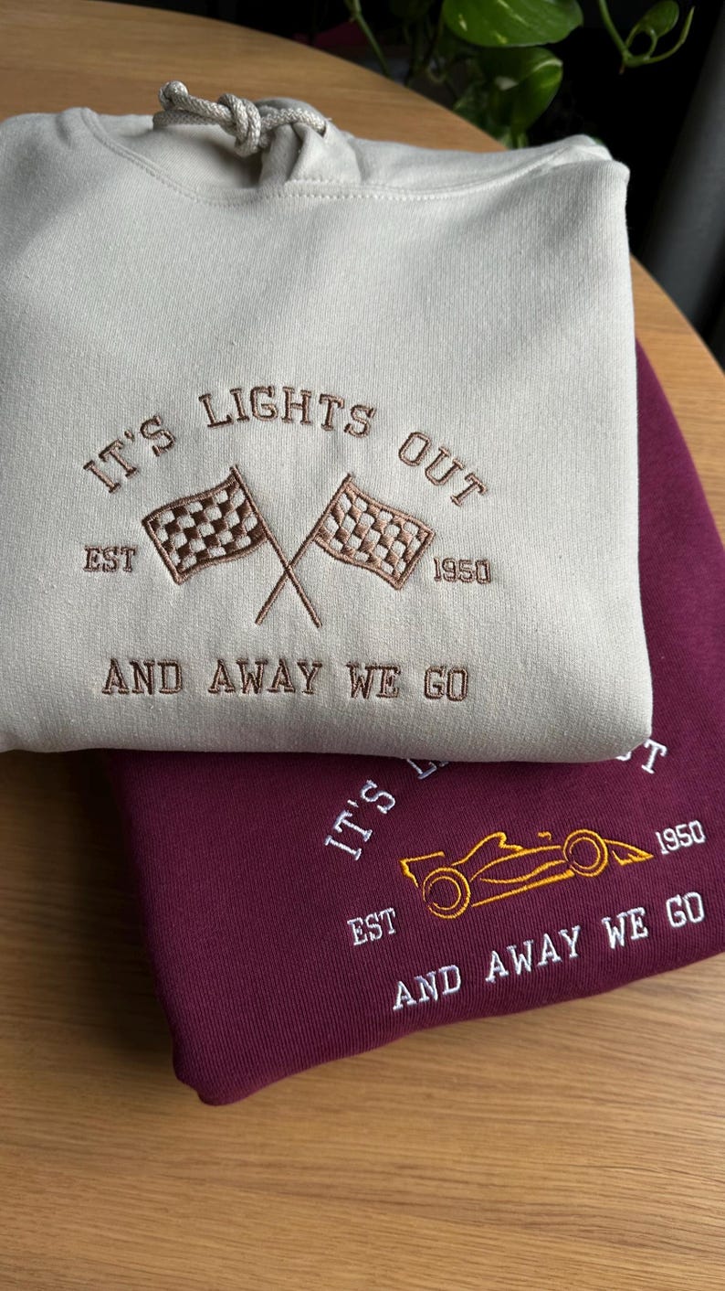 K&ouml;nnte beinhalten: Zwei Kapuzenpullover, einer beige und einer burgunderrot, mit gestickten Designs zum Thema Rennsport. Der beige Pullover hat ein kariertes Flaggen-Design und den Text "IT'S LIGHTS OUT EST 1950 AND AWAY WE GO". Der burgunderrote Pullover hat ein stilisiertes Rennwagen-Design und den Text "IT'S EST 1950 AND AWAY WE GO".