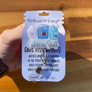 Könnte beinhalten: Ein Ausweishalter mit einem Cafeteria Staff Acryl-Aufsatz, der eine rosa Schürze, ein Tablett mit Pizza und Apfel sowie einen Milchkarton zeigt. Die Trägerkarte enthält Pflegehinweise und den Text Midwestern Design.