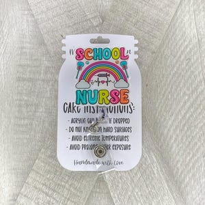 Op de afbeelding: Een witte kaart met een kleurrijke regenboog en de tekst "School Nurse" in een kleurrijk lettertype. De kaart bevat ook de tekst "Verzorgingsinstructies" en een lijst met instructies voor het verzorgen van de badgehouder. De badgehouder is gemaakt van helder acryl en heeft een zilveren intrekbare clip.