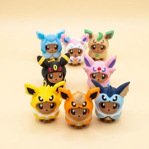 May include: A collection of eight small figurines featuring brown creatures wearing colourful hoods inspired by various characters. The hoods are blue, pink, tan, black, light pink, yellow, orange, and dark blue, each with distinct ear shapes.