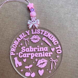 May include: A pink and white acrylic keychain with the text "Probably Listening to Sabrina Carpenter" and pink hearts, stars, lips, cherries, and a microphone.