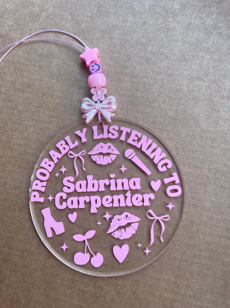May include: A pink and white acrylic keychain with the text "Probably Listening to Sabrina Carpenter" and pink hearts, stars, lips, cherries, and a microphone.