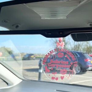 May include: A clear round car charm with the text "Probably Listening To Sabrina Carpenter" in pink lettering. The charm features pink hearts, cherries, a microphone, and a high heel shoe.