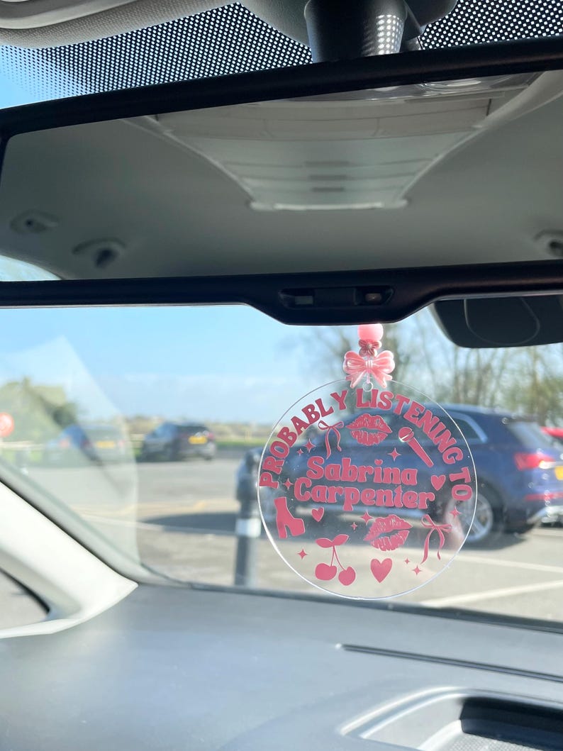 May include: A clear round car charm with the text "Probably Listening To Sabrina Carpenter" in pink lettering. The charm features pink hearts, cherries, a microphone, and a high heel shoe.