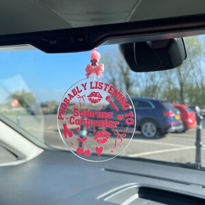 May include: A pink and red car charm with the text "Probably Listening To Sabrina Carpenter" and various pink and red icons including a microphone, a pair of high heels, a cherry, a heart, and a bow.