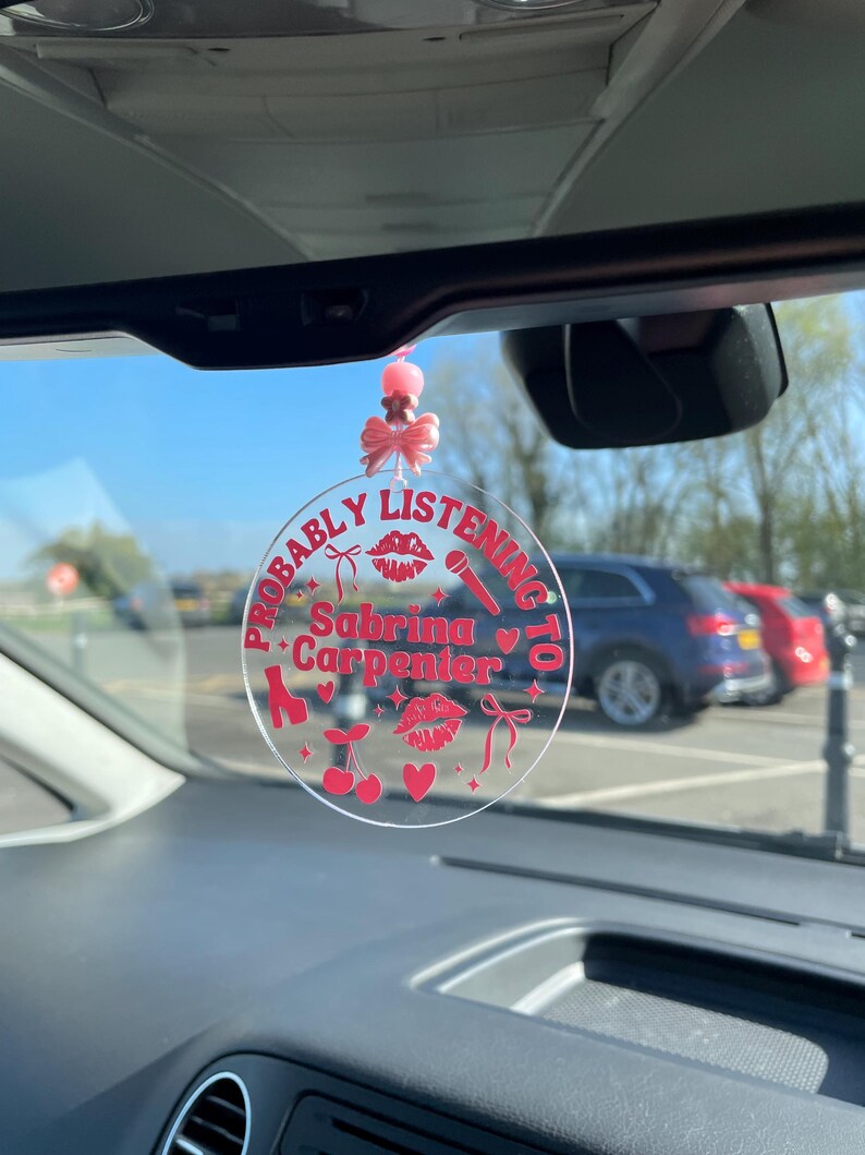 May include: A pink and red car charm with the text "Probably Listening To Sabrina Carpenter" and various pink and red icons including a microphone, a pair of high heels, a cherry, a heart, and a bow.