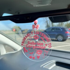 May include: A pink and white car charm with the text "Probably Listening to Sabrina Carpenter" and pink hearts, lips, bows, cherries, and a microphone.