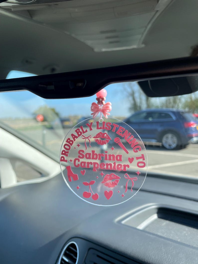 May include: A pink and white car charm with the text "Probably Listening to Sabrina Carpenter" and pink hearts, lips, bows, cherries, and a microphone.