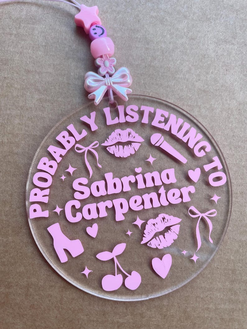May include: Pink acrylic keychain with the text "Probably Listening to Sabrina Carpenter" and pink hearts, lips, cherries, a shoe, and a microphone.