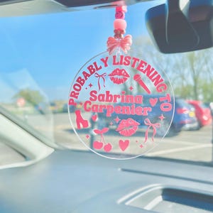 May include: A pink and white car charm with the text "Probably Listening to Sabrina Carpenter" and pink hearts, cherries, lips, and a microphone.