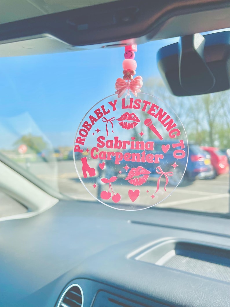May include: A pink and white car charm with the text "Probably Listening to Sabrina Carpenter" and pink hearts, cherries, lips, and a microphone.