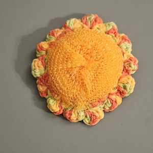Puede incluir: Un estropajo de cocina circular hecho a mano con un diseño de girasol. El centro es una malla de color naranja brillante, rodeada por un borde de ganchillo en tonos de naranja, verde y amarillo. El estropajo está diseñado para limpiar.