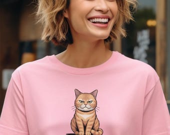 May include: Pale pink t-shirt with a cartoon cat illustration. The cat is depicted with a grumpy face and is brown and tan. The text below reads "MY CAT TELLS ME WHICH PEOPLE TO LIKE" in black capital letters.
