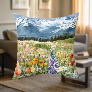 May include: Square throw pillow featuring a watercolor painting of a mountain range with snow-capped peaks. The foreground displays a vibrant meadow filled with colorful wildflowers in shades of pink, yellow, orange, and purple.