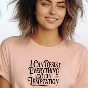 Puede incluir: Camiseta rosa claro con el texto negro "I Can Resist Everything Except Temptation". La camiseta tiene mangas cortas y cuello redondo. La persona de la foto está sonriendo.