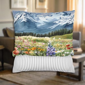 May include: A decorative pillow featuring a watercolor painting of a mountain range with snow-capped peaks. The foreground depicts a vibrant meadow filled with colorful wildflowers. The bottom pillow has a gray and white striped pattern.