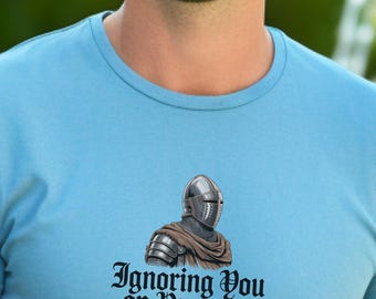 Könnte beinhalten: Hellblaues T-Shirt mit einer Grafik eines Ritters in Helm und Rüstung. Der Text "Ignoring You on Purpose" ist in einer dekorativen Schriftart unter der Ritterillustration aufgedruckt.