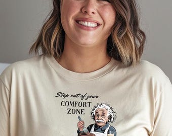 Op de afbeelding: Beige T-shirt met de tekst "Step out of your COMFORT ZONE" en een illustratie van Albert Einstein met een taart. De illustratie bevat een taart, aardbeien en een glas melk.