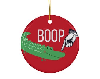 May include: Red round Christmas ornament with a green alligator and a pointing hand. The ornament says "BOOP" in white letters.