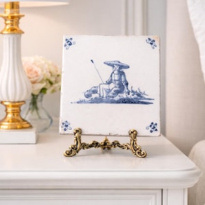 May include: A vintage blue and white ceramic tile featuring a seated figure with a wide-brimmed hat, holding a staff. The tile is displayed on a decorative gold-colored metal stand, showcasing a classic, detailed design. The tile is square.