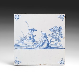 May include: Square ceramic tile with a blue and white pastoral scene. Two figures in hats are depicted near a tree and water, with a fishing rod. Decorative floral corner accents. The tile has a slightly aged appearance.