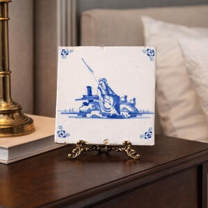 May include: A square, white ceramic tile with a blue illustration of a person fishing. The tile is displayed on a decorative brass stand. Floral corner accents frame the central image. The tile is on a dark wood nightstand.