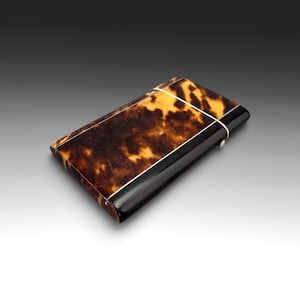 May include: A rectangular cigarette case featuring a mottled brown and black tortoiseshell pattern on the main body, accented by solid black end panels and thin silver-toned metal trim lines. A small silver-toned metal button is visible on the side.