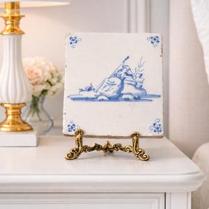 May include: A square, antique ceramic tile with a blue and white illustration of a classical scene, displayed on a decorative gold-colored stand. The tile has a distressed appearance and floral corner accents. The stand is ornate.