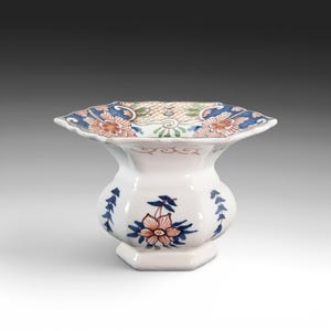 May include: White ceramic vase with a flared, scalloped rim. The rim features floral patterns in blue, orange, and green. The body of the vase is decorated with blue floral and leaf motifs. A decorative piece for the home.