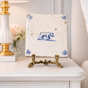 May include: A vintage, square ceramic tile with a blue bear illustration, displayed on a decorative gold-toned stand. The tile has blue floral corner accents. The scene is set on a white nightstand with a gold-based lamp and a vase of flowers.