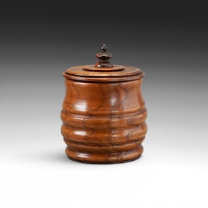 May include: A medium brown wooden jar with a smooth, polished finish and a cylindrical body featuring three horizontal, rounded ridges. The jar includes a matching circular lid topped with a small, pointed, dark brown finial handle.