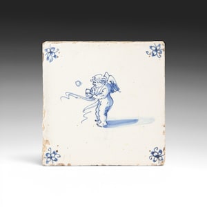 Puede incluir: Azulejo cerámico cuadrado con diseño pintado a mano. La imagen presenta un querubín azul y blanco sosteniendo una bola y una cinta. Acentos florales azules en cada esquina. El azulejo tiene una apariencia ligeramente envejecida.